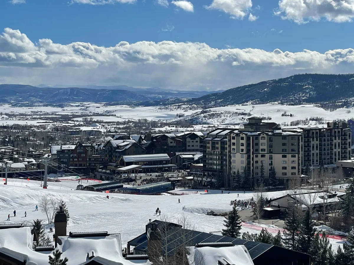 A bustling exterior view of resort buildings set against snowy slopes with skiers, a mountain backdrop, and vibrant après-ski activity. The scene is energetic, capturing both the excitement of the mountain and the lively social atmosphere at the base area.