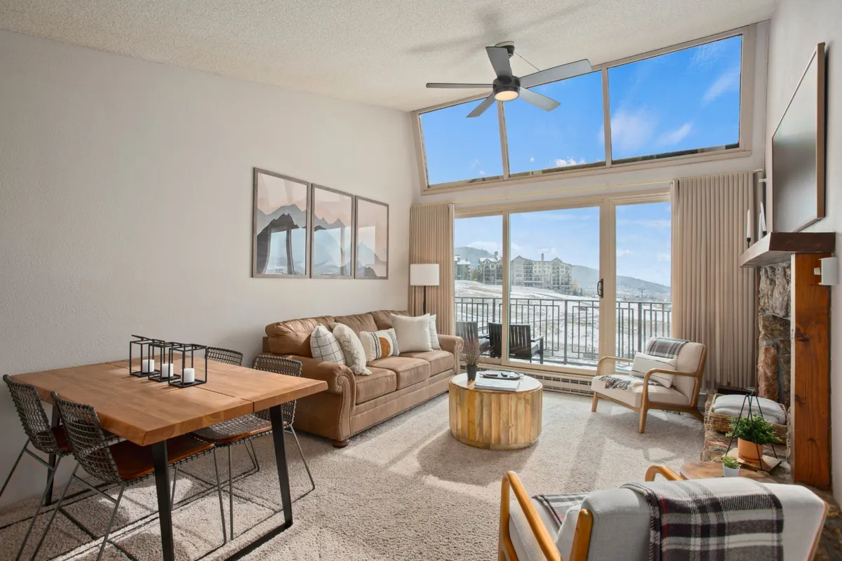 A spacious living room bathed in natural light from large windows, showcasing a comfortable brown sectional sofa, a modern dining table with industrial design, and a decorative round coffee table. Through the windows, scenic mountain views are visible, creating a seamless connection between the cozy interior and the dramatic alpine backdrop.