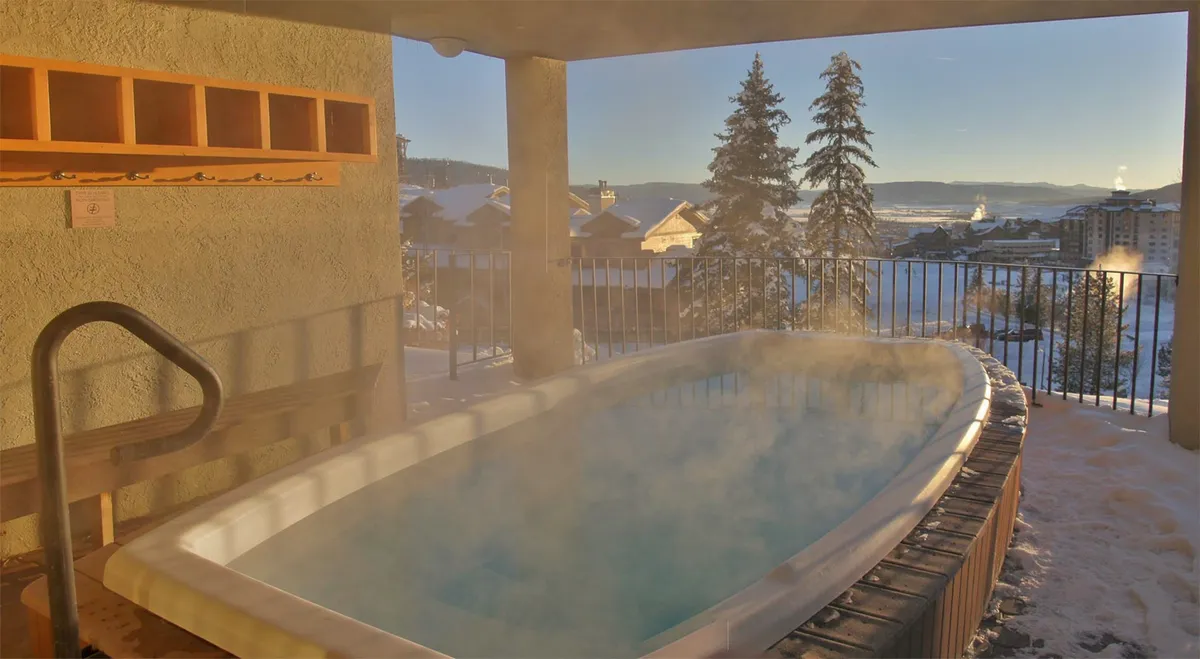 An outdoor hot tub set against a backdrop of snow-covered mountains and trees, creating an atmosphere of immersive relaxation. The warm steam rising from the tub contrasts with the crisp winter air, inviting guests to soak in the serene landscape.