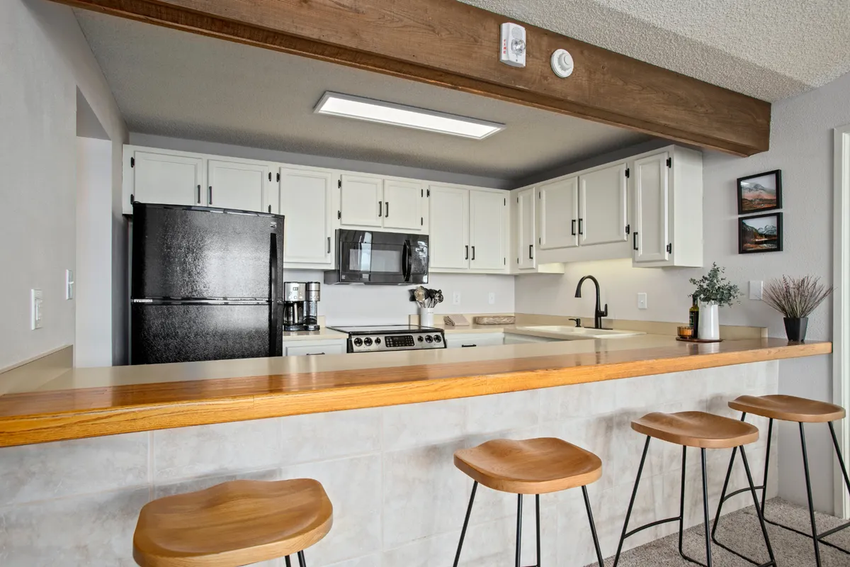 A stylish kitchen scene with a central island, multiple bar stools, modern white cabinetry, black appliances, and a wooden beam overhead. The layout is open and inviting, with plenty of space for cooking, dining, and entertaining guests.