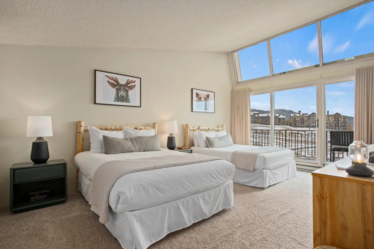 A welcoming double queen bedroom suite, with two neatly made beds featuring soft linens. Artwork adorns the walls, and large windows let in ample light while providing a glimpse of the alpine surroundings. The room feels inviting and ideal for families or friends.