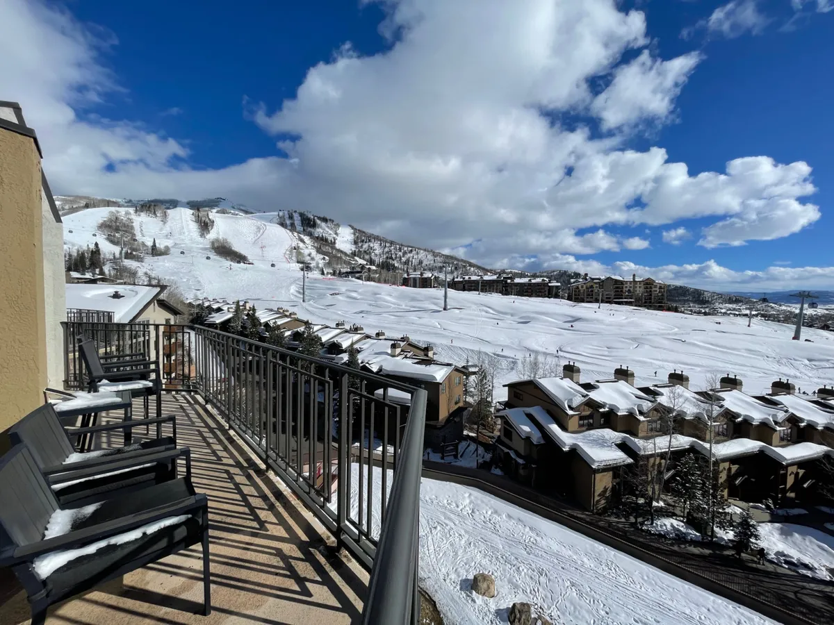 A private balcony with comfortable seating overlooks snow-covered slopes and a breathtaking mountain backdrop. Residential buildings and ski lifts dot the scenery, offering a direct visual link to the excitement of the ski base area.