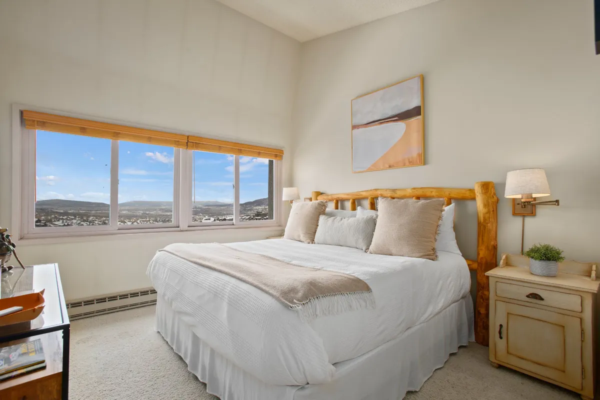 A serene king bedroom suite featuring a wooden bed frame adorned with soft, plush bedding. Large windows offer a scenic mountain view, filling the room with natural light. A nightstand with a lamp and a decorative art piece add upscale touches, evoking both comfort and style.