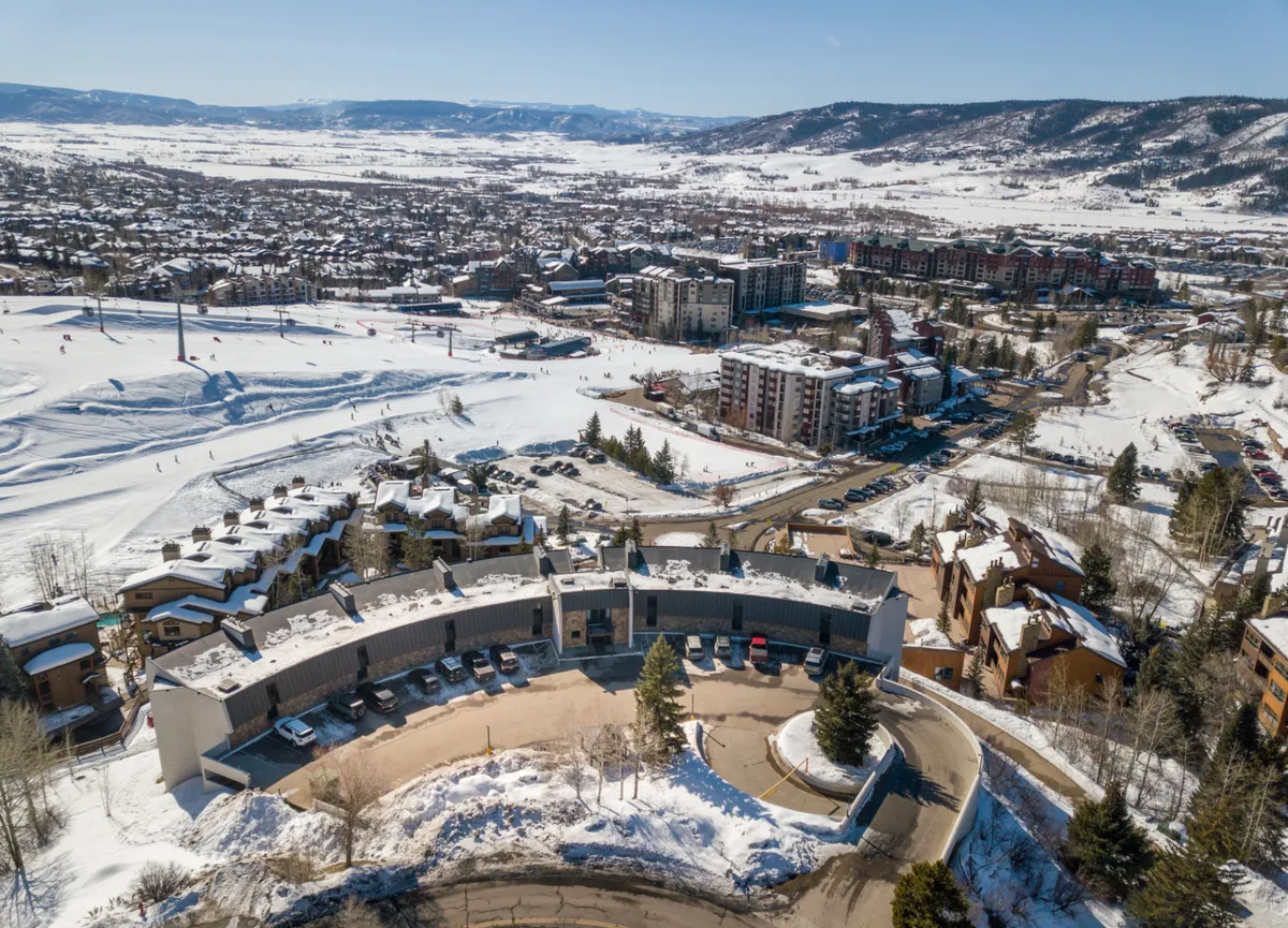 A lively exterior scene showcasing a modern building nestled at the base of snow-covered mountains, with ski slopes and residential areas nearby. The surroundings suggest easy access to both outdoor adventure and local amenities, with the structure suggesting proximity to shops and restaurants.