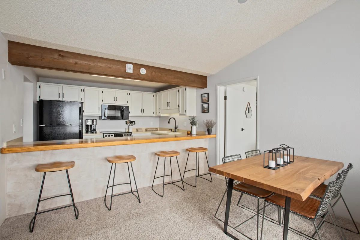 A bright, welcoming kitchen and dining nook are pictured with a central kitchen island, wooden dining table, and multiple bar stools. Kitchen appliances are visible, and the open layout encourages conversation between the cooking and dining spaces.